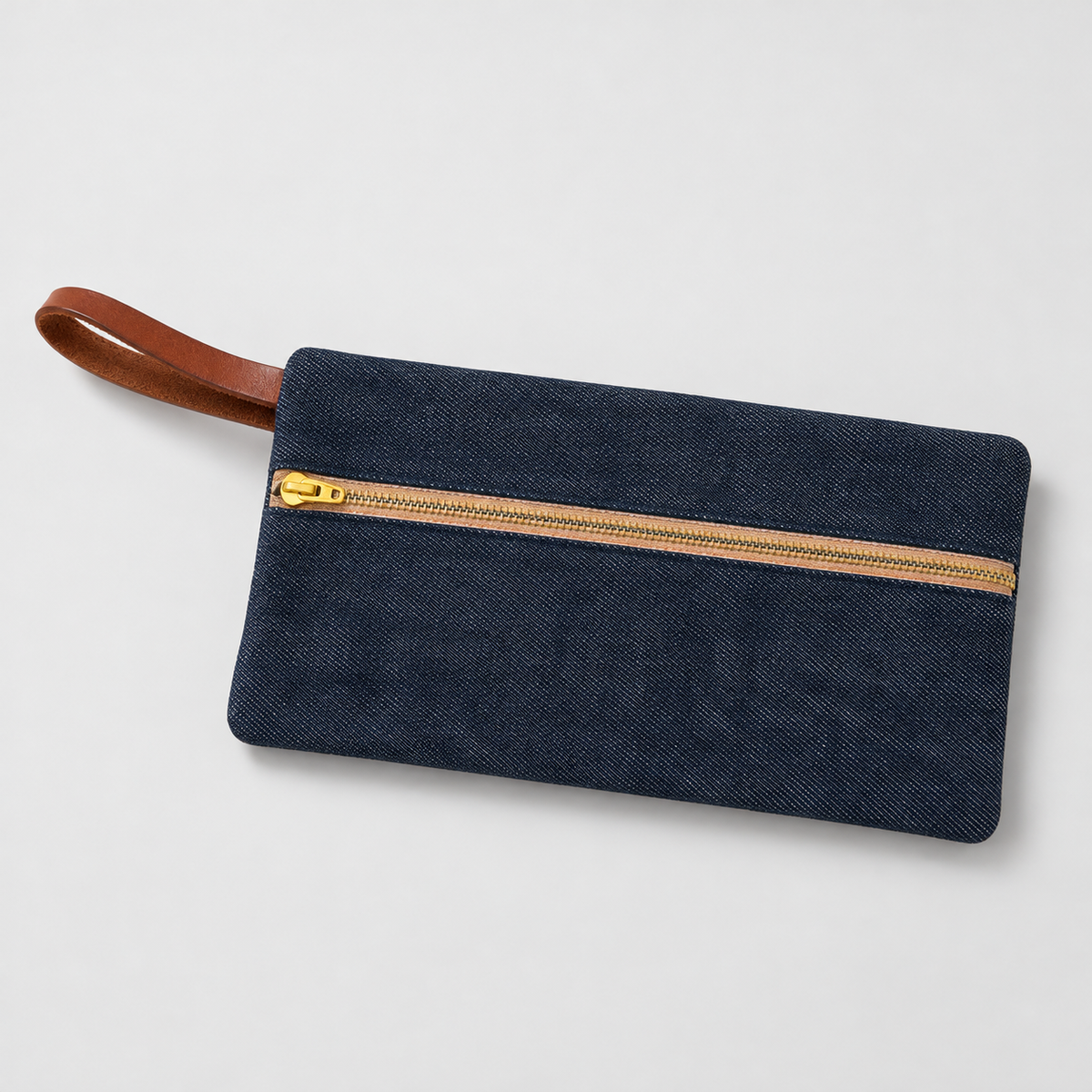 Pliegue 01 — Indigo — Indigo selvedge denim pouch with brass zipper and brown leather wrist loop, studio view